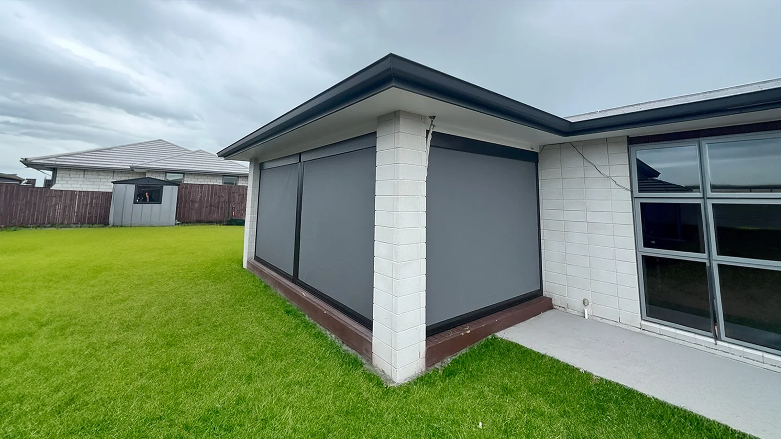 Front wide view of a modern dark charcoal track-guided outdoor blind installed on a backyard patio frame.